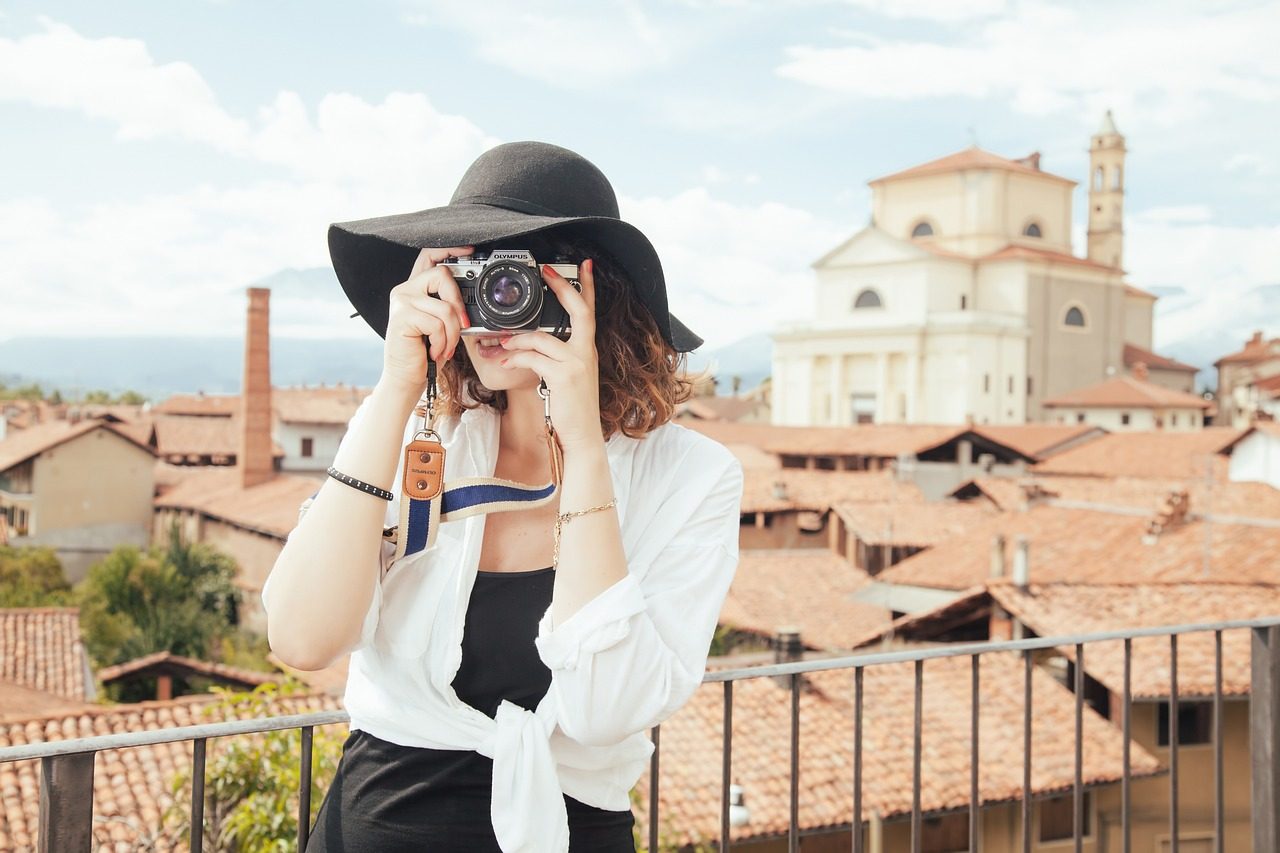 A woman in a black hate holding a camera and snapping a photo on a balcony somewhere in Italy
