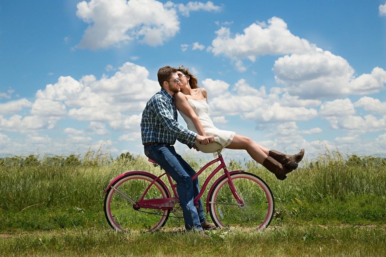 A romantic couple riding a bicycle through a field on a beautiful day