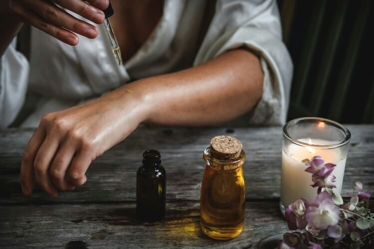 Woman with serum dropper dropping oil on her arm. She sits at a table with a candle, flowers, and DIY beauty products.
